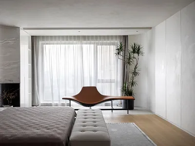 Minimalist bedroom with a wooden bed and a sleek, curved brown lounge chair in front of a large window with sheer curtains.