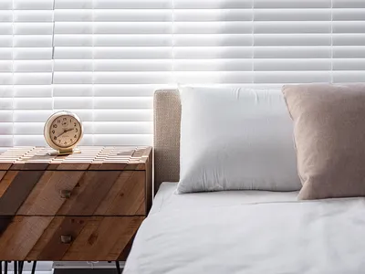 A minimalist bedroom featuring white blinds, a bed with white sheets and pillows, and a wooden side table with a clock.