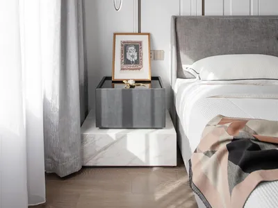 Modern minimalist bedroom featuring grey upholstered bed and marble nightstand with abstract artwork.