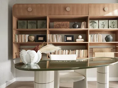 A modern home office featuring a wooden bookshelf filled with books and decorative items and a glass-top oval table with whit