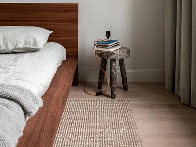 A minimalist bedroom featuring wooden furniture and a hanging light bulb on a stool, with a framed abstract painting above th
