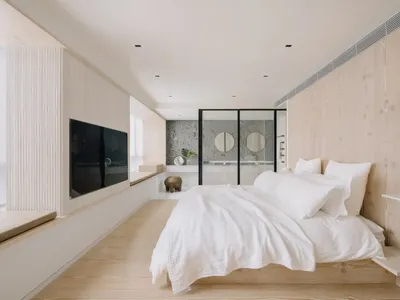 A minimalist bedroom with wooden flooring and a white bedspread, featuring a bathroom in the background with circular mirrors