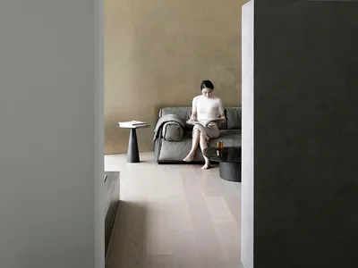A minimalist living room with a concrete wall, wooden floor, and a person reading a book on a sofa.