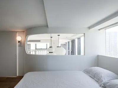 Modern minimalist bedroom with white walls, bed, and furniture, featuring a unique curved mirror and white lighting fixtures.