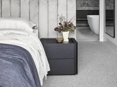 A modern bedroom featuring a gray wall with a textured pattern, a bed with white bedding, and a dark nightstand with a plant.