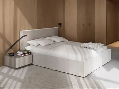 A minimalist bedroom with a white bed sheet, a headboard, and a nightstand with a lamp and books.