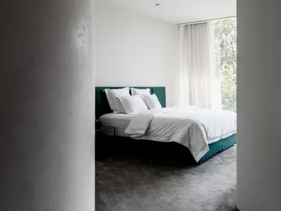 A minimalist bedroom with concrete walls and a bed with a green headboard and white bedding.