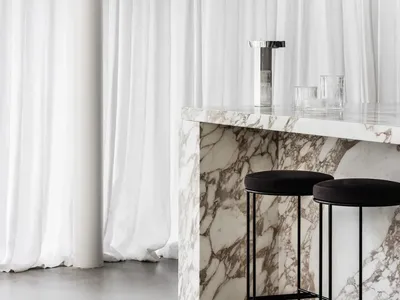Bar stools placed at a marble counter in front of white curtains