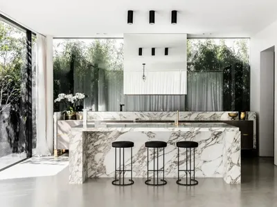 Modern kitchen featuring marble countertops and stools, with large windows showcasing greenery.