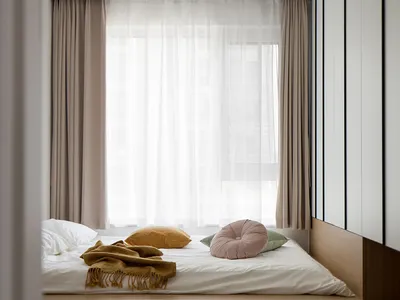 A minimalist bedroom featuring a low wooden bed with soft cushions and a blanket, under large windows with sheer curtains.
