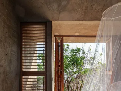 A minimalist bedroom with concrete flooring, wooden shutters, and a large bed covered in a white curtain.