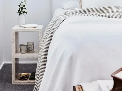 Minimalist bedroom with white walls, white bedding, and a small nightstand with greenery and candles.