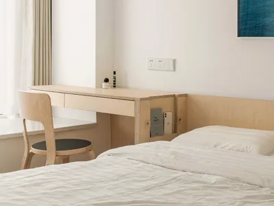 A minimalist bedroom featuring a wooden desk and chair, a bed with white bedding, and a window with light-colored curtains.