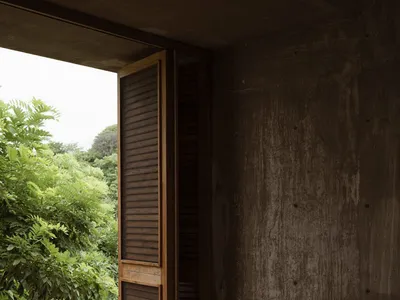 A modern bedroom featuring a concrete wall and a macrame chair, with a view of lush greenery outside through an open window.