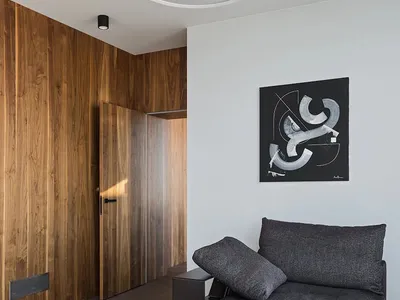 A minimalist living room featuring a wooden wall, glass coffee table, and a grey armless sofa with a mounted artwork.