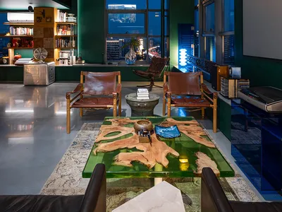 A modern industrial living room featuring glass walls, leather chairs, metallic accents, and wooden elements.