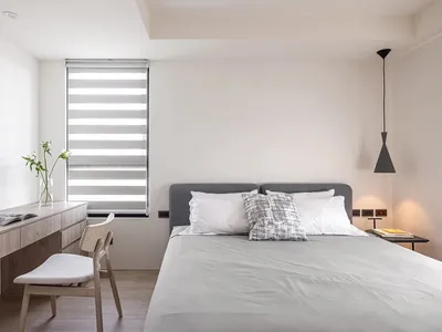 A minimalist bedroom with a fabric bed, wooden desk, and roller blinds.