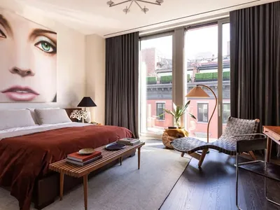 Modern bedroom with a large wall mural of a woman's face, wooden bed frame, fabric bedding, metal floor lamp, and wooden benc