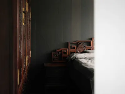 A minimalist bedroom with a wooden headboard, neatly made bed, and dark wooden furniture against a dark grey wall.
