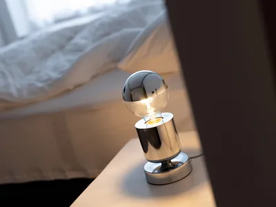 A minimalist bedroom featuring a metallic table lamp with a glowing lightbulb and a neatly made bed with white bedding.