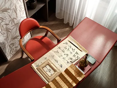 A modern home office with a red leather desk and chair, featuring calligraphy and a traditional vase.