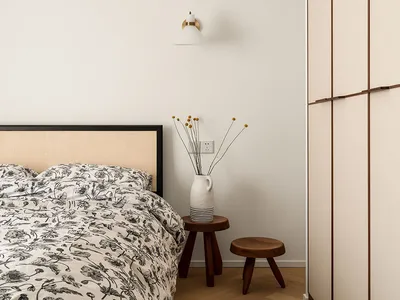 A minimalist bedroom with a wooden headboard bed, floral bedding, and a simple wooden stool with a vase of flowers.
