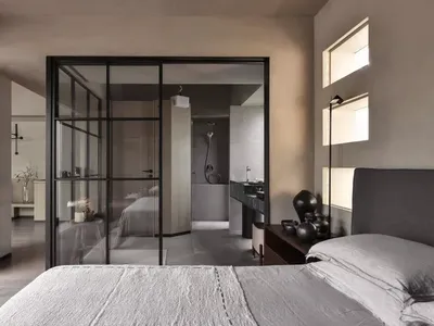 A minimalist bedroom with a concrete floor and walls, featuring a large bed and a glass-enclosed bathroom in the background.