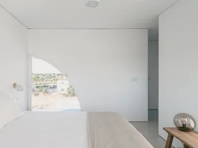 Modern minimalist bedroom with concrete walls and floor, featuring a large curved window overlooking a natural landscape.