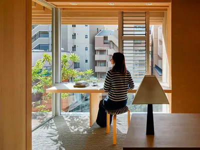 A person sitting at a wooden desk in a home office, with a chair and a lamp on a wooden table.