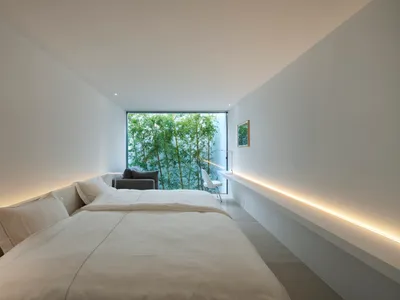 Modern, minimalist bedroom with white walls, glass window overlooking greenery, and a simple, comfortable bed.