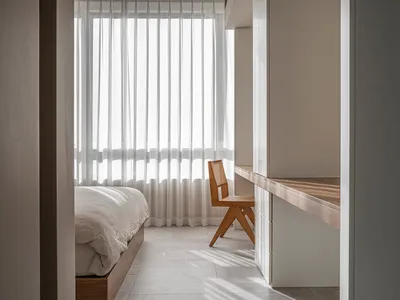 Minimalist bedroom featuring a bed with white sheets and wooden chair against a white background with vertical white curtains