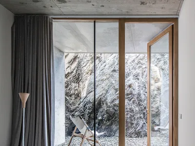 A modern minimalist bedroom with a concrete floor and ceiling, featuring a large window with a view of rocky terrain, a woode