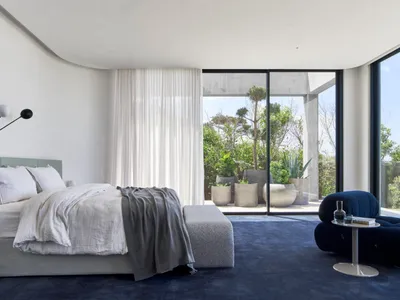 A minimalist bedroom featuring fabric bedding, glass and concrete surfaces, and a large window with a view of greenery outsid