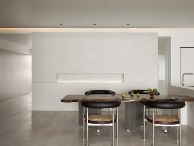 A modern minimalist kitchen with a wooden table and minimalist chairs in front of a concrete wall with recessed lighting. The