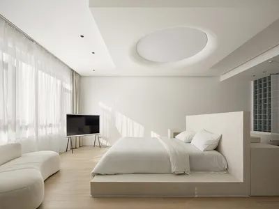 A minimalist bedroom featuring white furniture and a large window with sheer curtains.