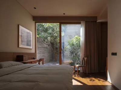 A serene bedroom with a wooden floor, a bed with a wooden headboard, and a large window or sliding glass door leading to a co