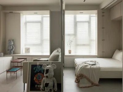 A minimalist bedroom with a bed, couch, and window with blinds. Room features a neutral color palette and simple furnishings.