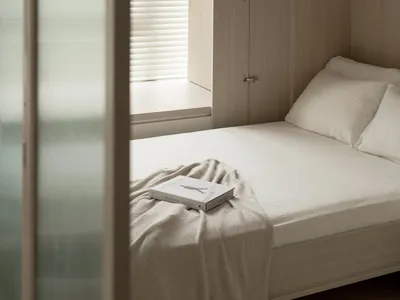 A minimalist bedroom with a white bed featuring a book on the bedspread, surrounded by neutral tones and soft lighting.