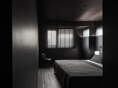 Minimalist bedroom with wood flooring and furniture, featuring a single window with blinds and a lit lightbulb.
