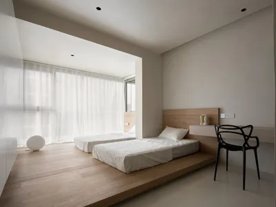 A minimalist bedroom featuring twin beds on a wooden platform with sheer curtains and a black chair against a white wall.
