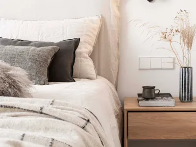 A modern minimalist bedroom with a bed featuring neutral-colored towels and a wooden nightstand with a vase of dried grasses