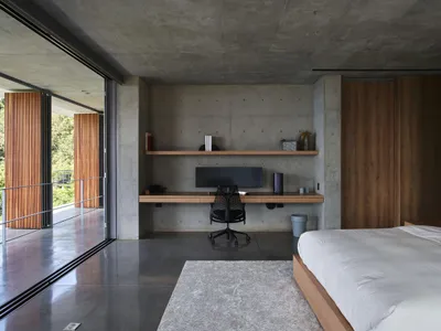 A minimalist bedroom with concrete floors, walls, and furniture, featuring a large bed, a floating desk, and a built-in shelf