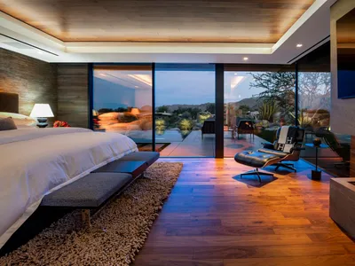 Modern bedroom featuring brown wood flooring, large windows with outdoor view, and a cozy seating area.
