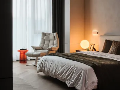 A minimalist bedroom with white and blush tones, featuring a modern armchair, a cozy bed with a textured blanket, and a warm