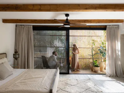 A modern minimalist bedroom with a wooden ceiling, a bed covered in beige linen, and potted plants in the background.