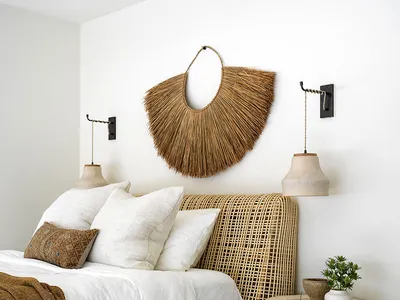 A minimalist bedroom featuring a wicker bed with a white comforter and pillows, a woven basket wall decoration, and a wooden