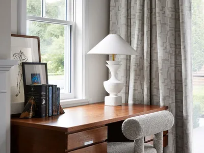 A modern home office featuring a wooden desk, fabric-upholstered chair, and glass-paneled window with patterned curtains.