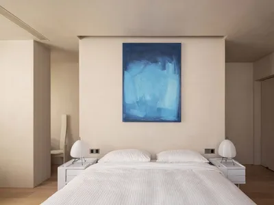 Modern minimalist bedroom with a blue abstract painting above the bed, white bedding, and two cylindrical lamps on side table