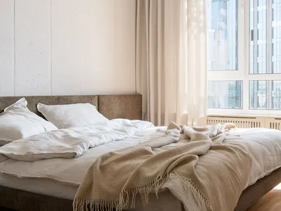 A minimalist bedroom with neutral-toned bedding and curtains, featuring a wooden floor and a concrete-style white wall.