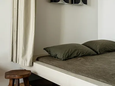 A minimalist bedroom featuring a bed with a wooden headboard and green bedding, alongside a wooden stool and a curtain.